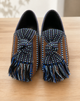 A pair of slip-on loafers with woven brown side panels, blue and white patterned uppers, and long fringe tassels, displayed on a light wood surface.