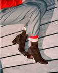 A close-up of a person sitting on weathered gray wooden planks, wearing dark brown suede ankle boots with low block heels, light gray trousers, and a bright red top visible at the top edge.
