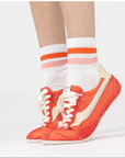 Orange sneakers with white laces worn with white socks featuring red stripes on a light gray background