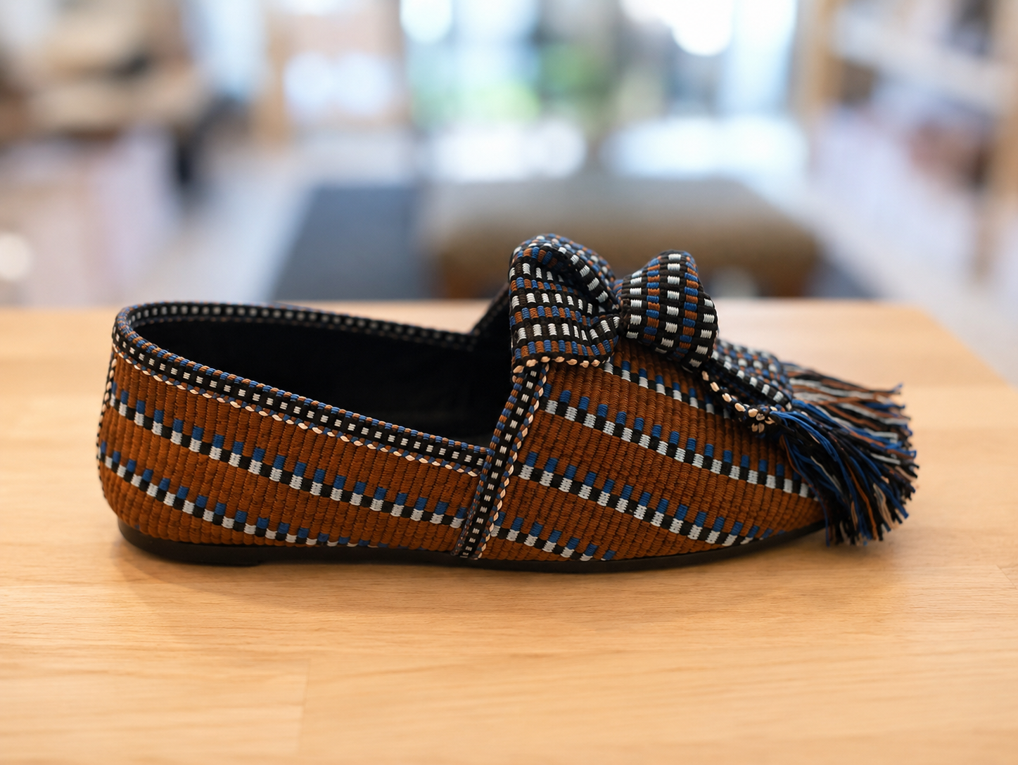 A single woven loafer-style slipper with a bow and tassel, shown in side profile on a light wood surface. The upper is primarily brown with woven blue, white, and black geometric stripes, and the bow and tassel are black with blue and white woven accents.