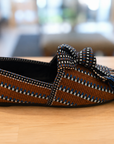 A single woven loafer-style slipper with a bow and tassel, shown in side profile on a light wood surface. The upper is primarily brown with woven blue, white, and black geometric stripes, and the bow and tassel are black with blue and white woven accents.