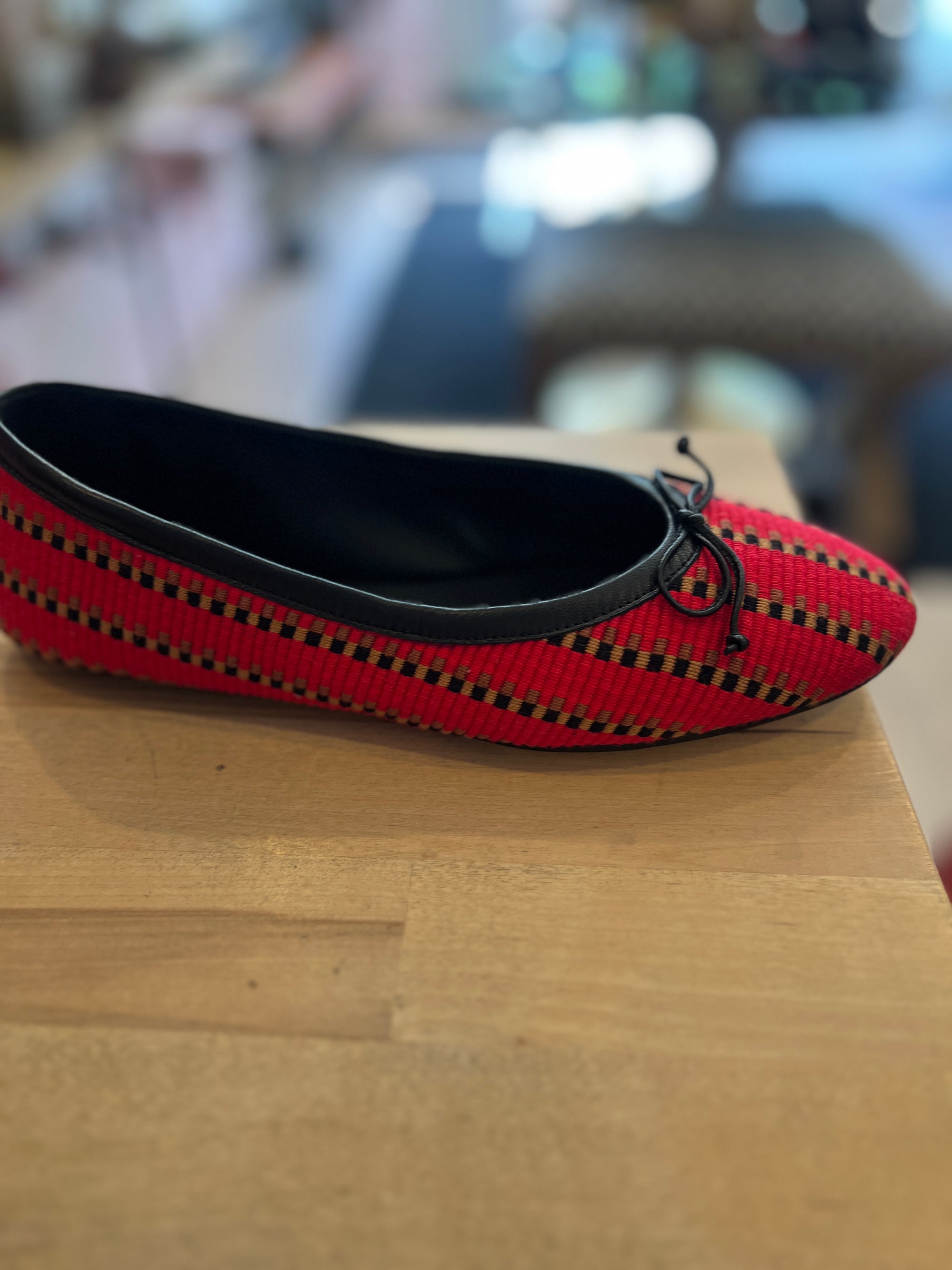 A red woven ballet flat shoe with black trim and a small black bow, resting on a light wood surface with a blurred indoor background.