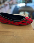 A red woven ballet flat shoe with black trim and a small black bow, resting on a light wood surface with a blurred indoor background.