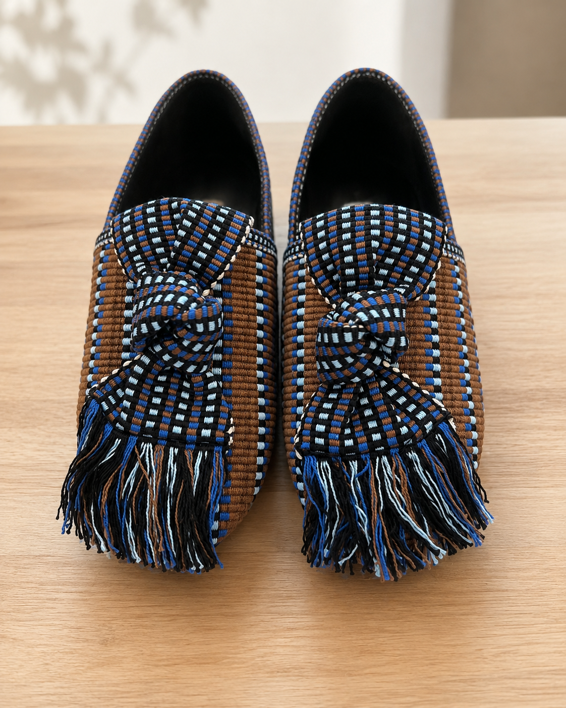 A pair of slip-on loafers with woven brown side panels, blue and white patterned uppers, and long fringe tassels, displayed on a light wood surface.