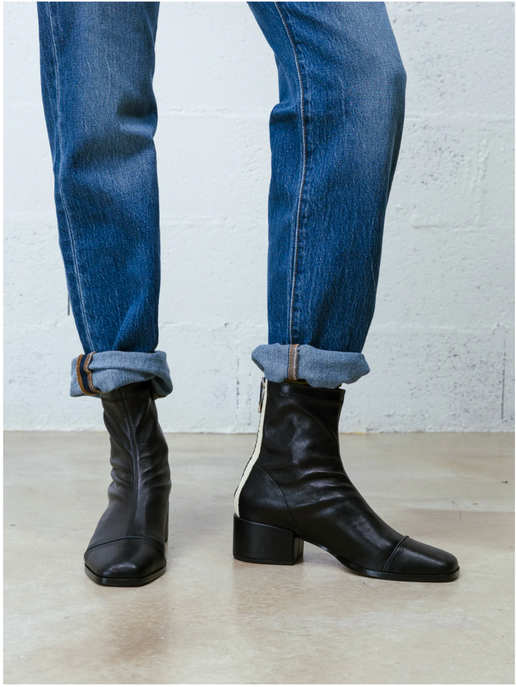 Black leather ankle boots with a block heel and side zipper, worn with cuffed blue jeans against a white brick wall and concrete floor.