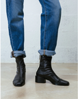 Black leather ankle boots with a block heel and side zipper, worn with cuffed blue jeans against a white brick wall and concrete floor.
