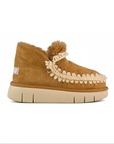 A single tan suede moccasin-style slipper with a cream braided trim, fluffy pom-pom, and thick cream sole, shown in profile against a white background.