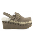 A taupe-brown suede clog shoe with a white shearling lining, a platform sole, and bronze-tone metal studs and buckle details, shown from a side profile on a white background.