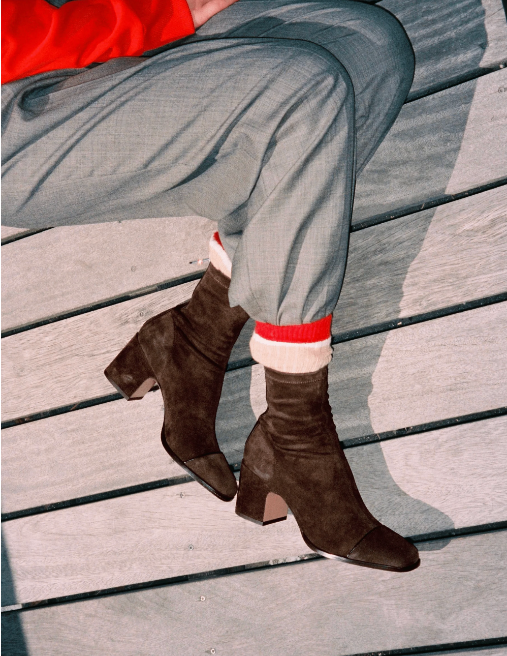 A close-up of a person sitting on weathered gray wooden planks, wearing dark brown suede ankle boots with low block heels, light gray trousers, and a bright red top visible at the top edge.