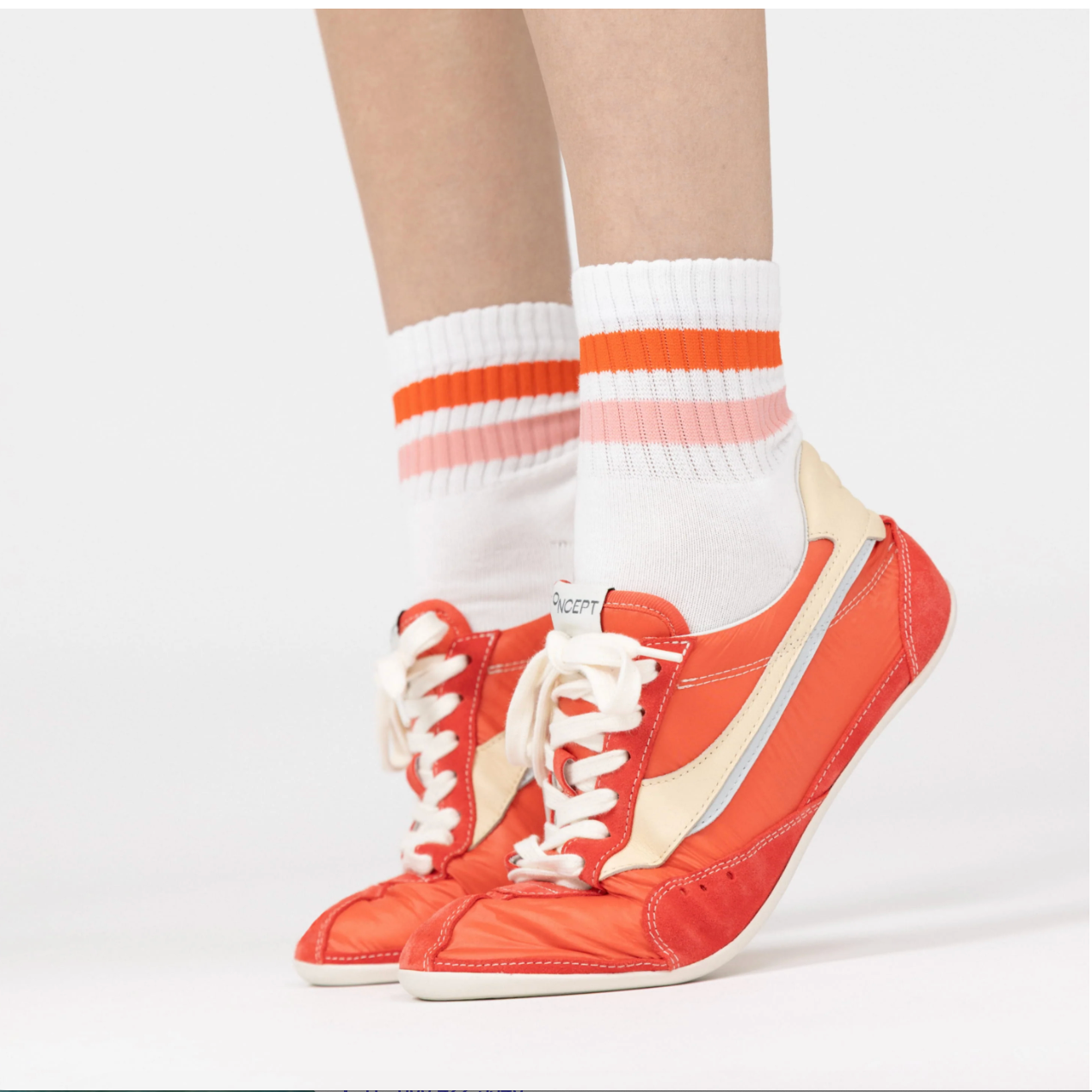Orange sneakers with white laces worn with white socks featuring red stripes on a light gray background